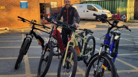 Man stands with four e-bikes in a row. Bick building a white van in background.