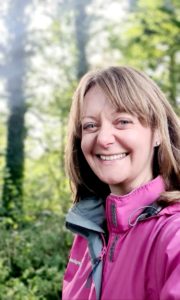 Katherine smiles at camera. Wearing pink jacket, tall green leafy trees in the background.