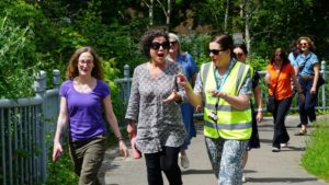 Three woman walk at the front of a walking group, chatting and smiling, green trees and path in the background. Woman on the right hand side wears yellow high viz vest.