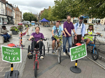 Five people in a row astride and seated on bikes/ adapted tricycles and scooters. Green signs in front of the group read: 'Road open for' and images of people walking, wheeling and cycling are shown.