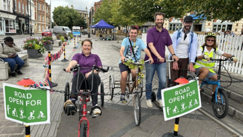 Five people in a row astride and seated on bikes/ adapted tricycles and scooters. Green signs in front of the group read: 'Road open for' and images of people walking, wheeling and cycling are shown.