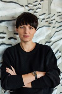 Woman stands with arms folded. She has dark cropped hair and wears a black jumper.