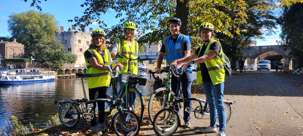 Four people stand by river holding bikes. They wear cycling helmets and high vis vests.