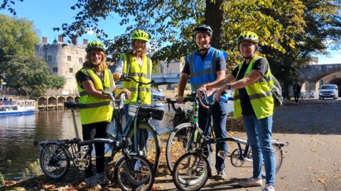 Four people stand by river holding bikes. They wear cycling helmets and high vis vests.