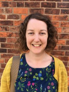 Woman smiles at camera. She has curly brown hair and wears a yellow cardigan over a floral print blue top