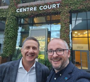 Two men pose outside a building. Sign on the building above large glass door reads: Centre Court. Building has leafy green coverage over brickwork.