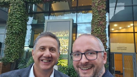 Two men pose outside a building. Sign on the building above large glass door reads: Centre Court. Building has leafy green coverage over brickwork.