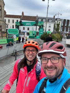 Man and woman pose for camera wearing cycle helmets. The woman on the left wears a bight pink cycle jacket and the man in the right a bright blue cycle jacket.