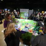 A busy exhibition hall. People looking at display stands and chatting. In the foreground two woman look at brightly coloured leaflets with 'Brightwayz' logo on.