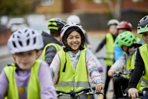 Image credit: Bikeability. Group of children smiling and laughing riding bikes. They wear cycle helmets and yellow high viz vests. 