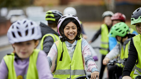 Image credit: Bikeability. Group of children smiling and laughing riding bikes. They wear cycle helmets and yellow high viz vests.