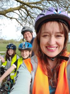 Image credit: Bikeability. Emily Cherry poses for the camera, wearing cycle helmet and orange high viz vest. Her family - two children and a man, are behind her. They each wear a cycle helmet and high viz vest. A tree and green field can be seen in the background.