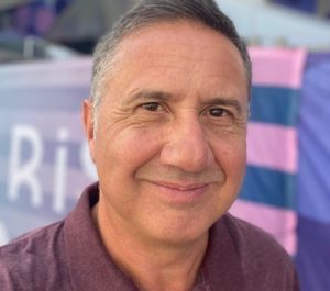 Man smiles at the camera. He's wearing a purple polo neck top.