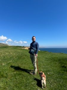 Man stands on green grass with small brown dog on a lead. The sky is blue, with small white clouds and a cliff edge can been seen behind him. The dark blue sea is in the background. 