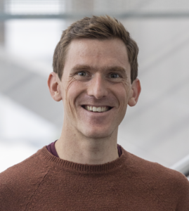 Head and shoulder shot of man smiling at camera. He wears light brown jumper. Background is white.