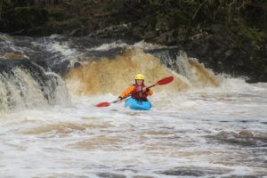 Woman smiles, exhilarated as she kayaks down a foamy river, water fall in background. She wears a yellow helmet and red inflatable jacket.