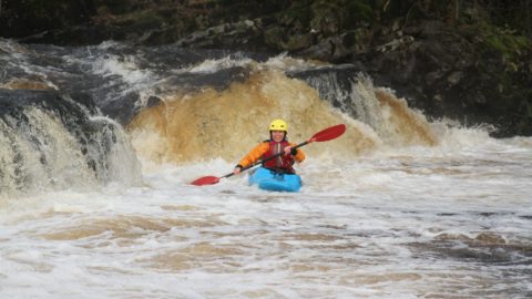 Woman smiles, exhilarated as she kayaks down a foamy river, water fall in background. She wears a yellow helmet and red inflatable jacket.