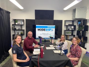 Four people sit in a studio set. There are microphones on the table in front of them. A Modeshift logo is on a large screen in the background.