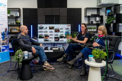 Three people sit either side of a large screen with video conference call in progress. They are in a studio surrounded by microphones.