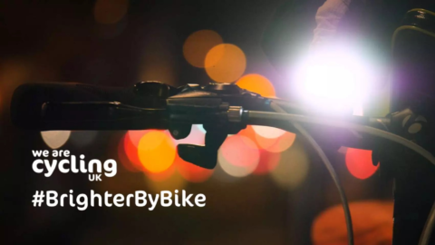 It's night time, a bike handle bars with red lights out of focus in the background. Text reads: We are Cycling Uk. #BrighterByBike