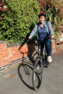 Jenny sits astride her bike. She wears a cycle helmet, blue trousers and top with grey jacket. She smiles at the camera. Pavement and redbrick wall, with green shrubbery next to her.