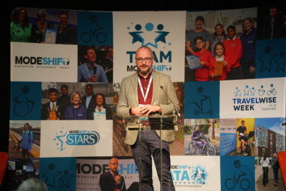 Man stands at lectern on stage. Modeshift branded display stand behind him.