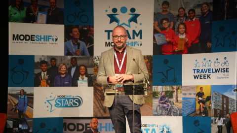 Man stands at lectern on stage. Modeshift branded display stand behind him.