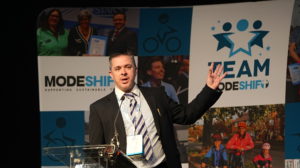 Man stands at lectern, gesturing with left hand. He wears a dark suit and striped tie.