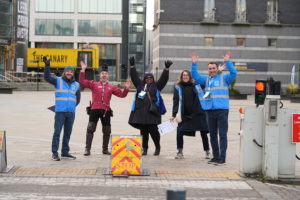 Group of people smiling and waving at the camera. In front of a building. They are wearing winter coats and bright blue Modeshift branded high viz vets.