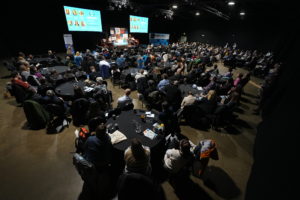 Convention room with low lighting. Delegates sit around round tables looking towards a stage with two large screens.