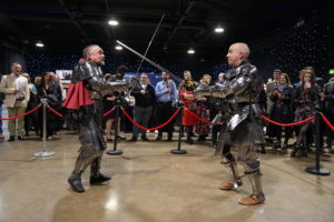 Two men in shining armor brandish swords, positioned prominently in front of a captivated audience, ready for action.