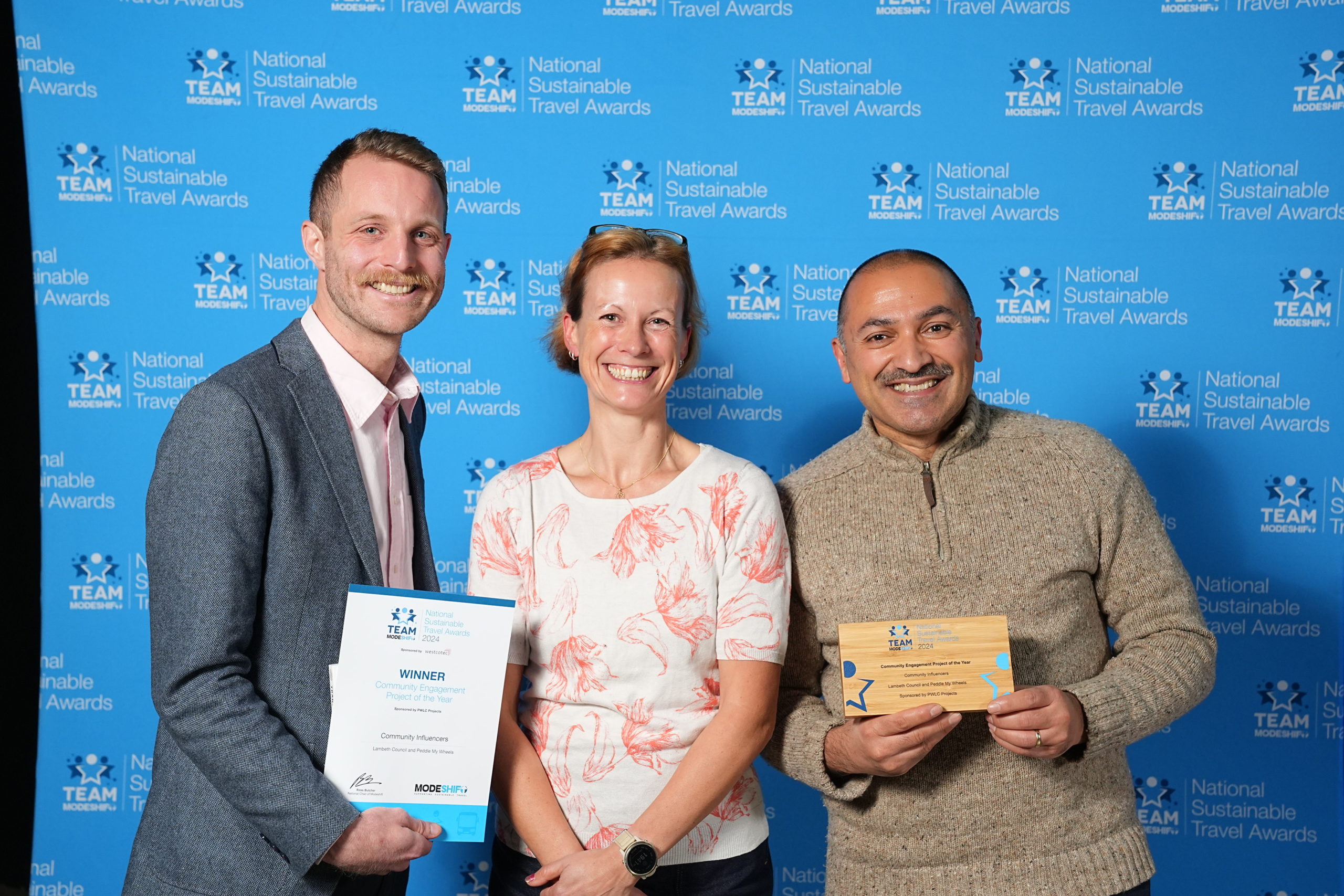 Three people stand together. Two men and a woman in the middle. The two men each hold a certificate and award trophy.