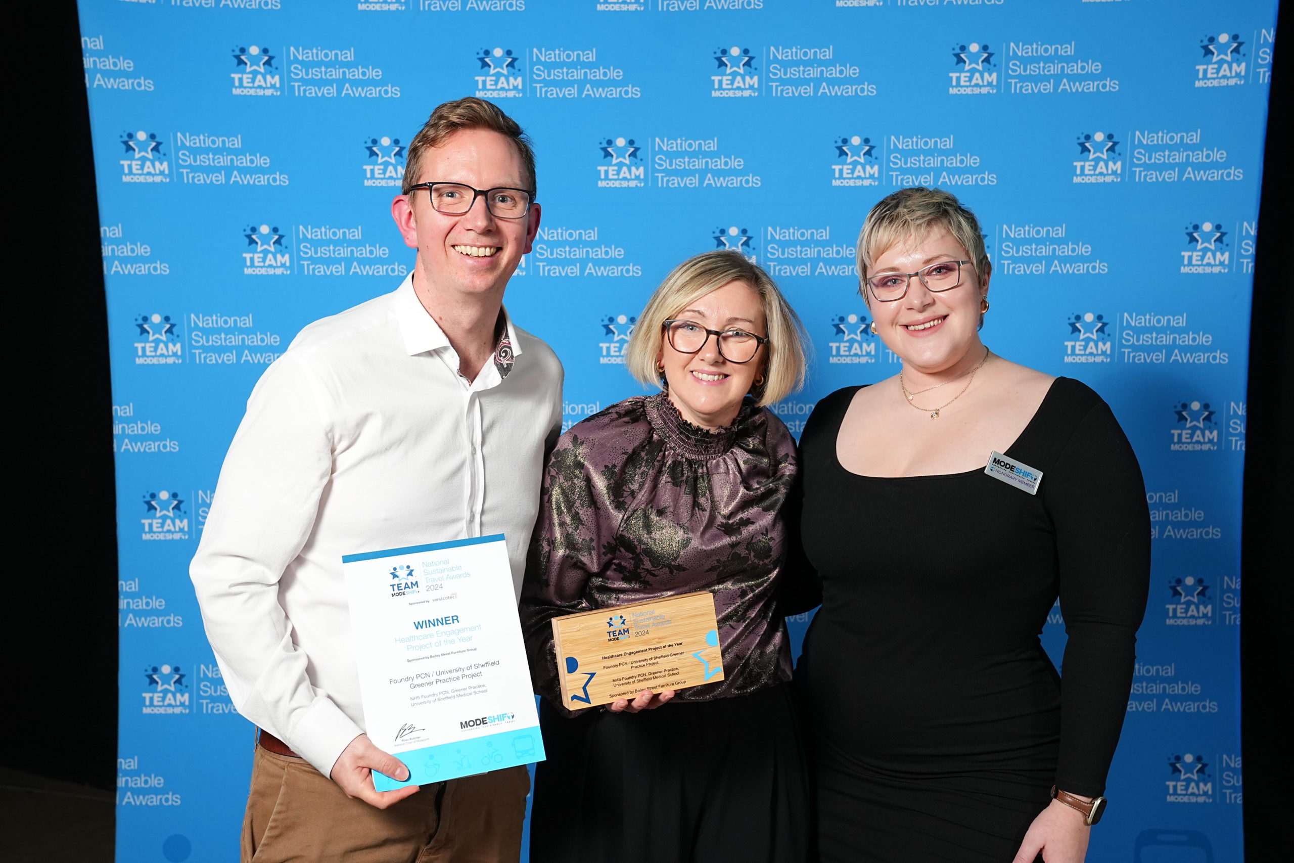 Three people stand together. The man and woman in the centre hold a certificate and award trophy.