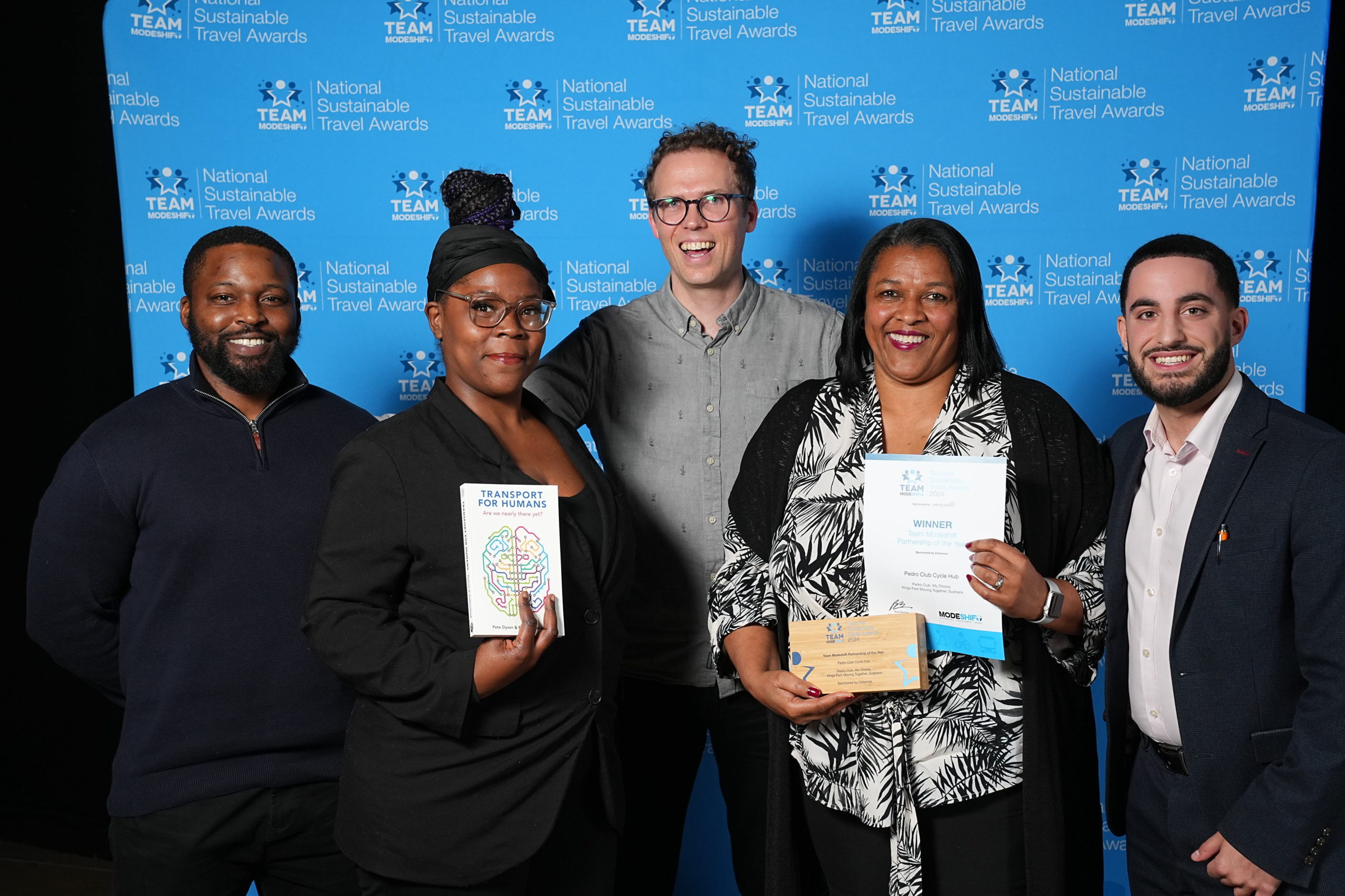 Five people stand together. The two women in the centre each hold a certificate and award trophy.