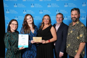 Five people stand together. Three women on the left hold award trophy and certificate.