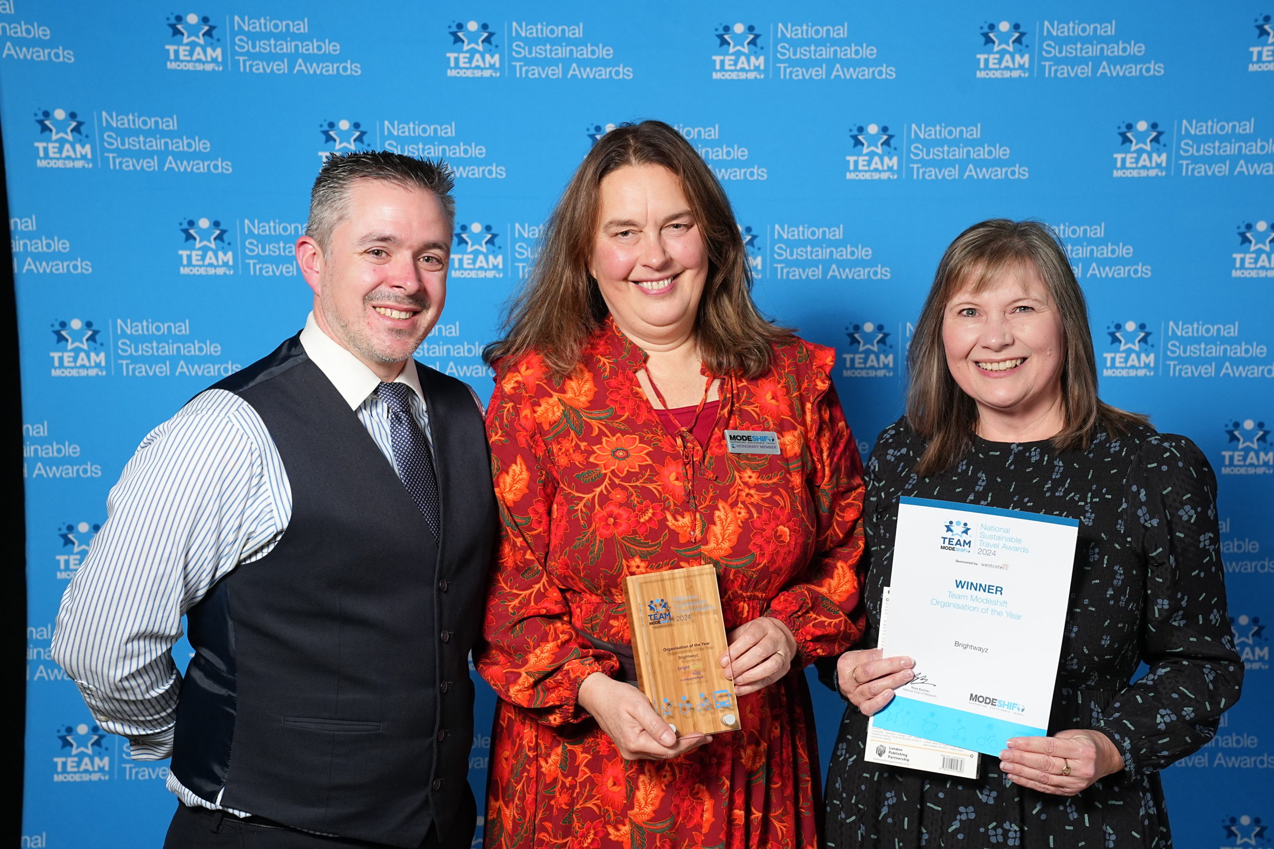 Three people stand together. Two women each hold a certificate and award trophy.