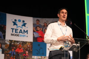 A man addresses an audience at a podium, with a "Modeshift" banner displayed prominently behind him.