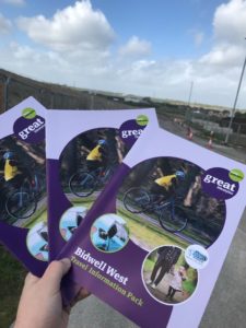 Hand holding three booklets in a fan. Purple booklets with a cyclist and text reads: 'Bidwell West Travel Information Pack'.