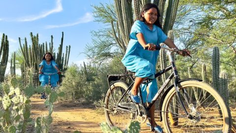 Two people in blue outfits ride bicycles on a dirt path through a desert landscape with cacti and shrubs under a blue sky. Credit World Bicycle Relief