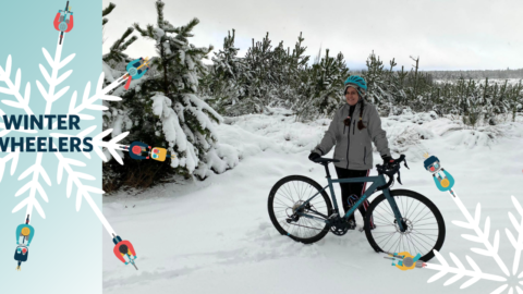 Snowy scene, woman stands by bicycle. Text on left hand side reads: 'Winter Wheelers'.