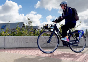 Cyclist peddles past a wall with 'Linmere' etched into it. Houses in the background.