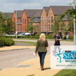 Two people walking down a paved residential street with houses and cars in the background. Greenery lines the street. Modeshift STARS Residential logo in teh bottom right corner. Text reads: 'Excellent Travel Plan 2024'.