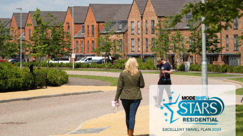 Two people walking down a paved residential street with houses and cars in the background. Greenery lines the street. Modeshift STARS Residential logo in teh bottom right corner. Text reads: 'Excellent Travel Plan 2024'.