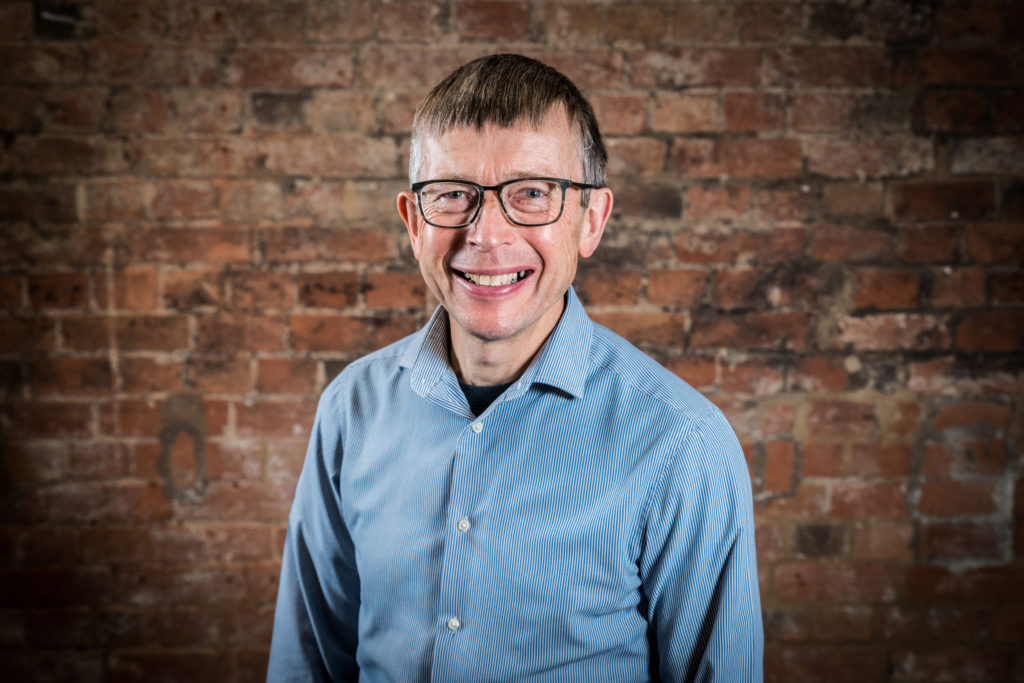 Man smiles at the camera. He wear a light blue shirt and glasses.