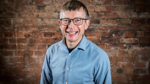 Man smiles at the camera. He wear a light blue shirt and glasses.