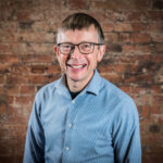 Man smiles at the camera. He wear a light blue shirt and glasses.