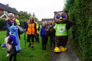 Group of school children in uniform walk with adults across grass/pathed area. They walk with a large owl mascot wearing a green t-shirt that reads: Leeds School Streets