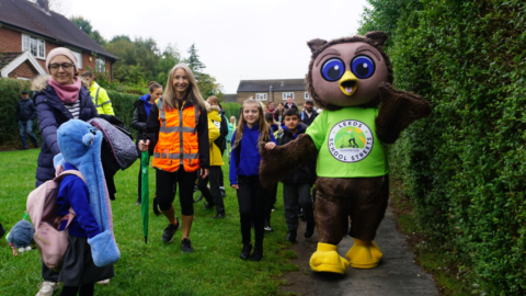 Group of school children in uniform walk with adults across grass/pathed area. They walk with a large owl mascot wearing a green t-shirt that reads: Leeds School Streets