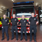 Four men in black t-shirts and trousers stand in a line in front of a fire engine.