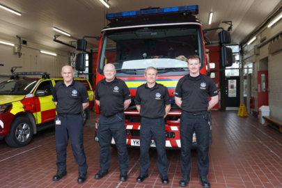 Four men in black t-shirts and trousers stand in a line in front of a fire engine.